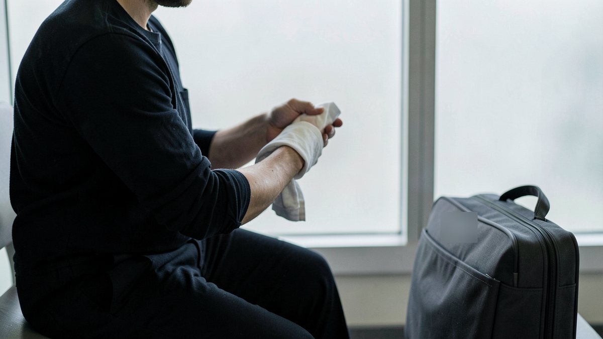 An adult seen from the chest down, seated on a terminal bench and holding a folded cold compress against their forearm, with a dark carry-on bag beside them